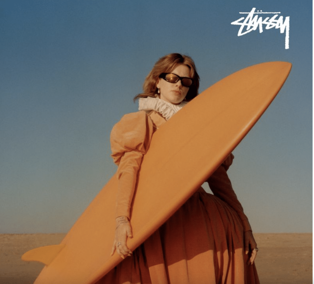 Model poses in Stussy dress with a surfboard on the beach